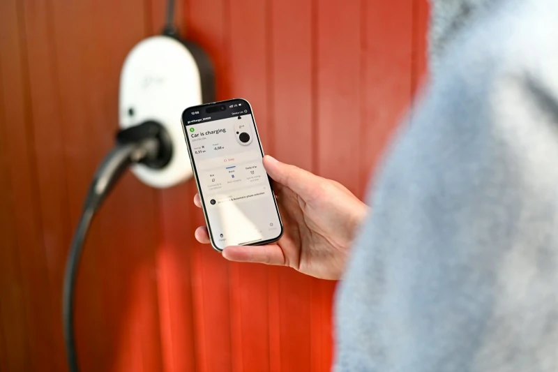 Iemand bekijkt een laadapp op de telefoon naast een witte laadpaal aan een rode gevel